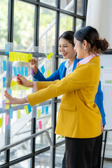 Strategic Collaboration: Two smiling businesswomen brainstorm ideas on a glass board using colorful sticky notes, fostering innovative teamwork in a modern office setting. 