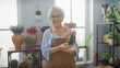 © Krakenimages.com - Smiling senior woman in apron holding orchid at a flower shop surrounded by various plants.