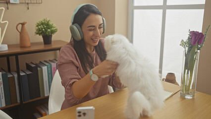 A cheerful hispanic woman enjoys time with her bichon maltese dog in a cozy living room, exemplifying a happy indoor lifestyle moment.