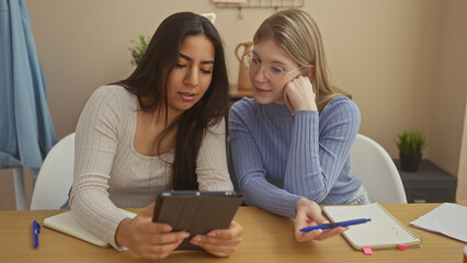 Two women studying together in a modern apartment, sharing a tablet and taking notes