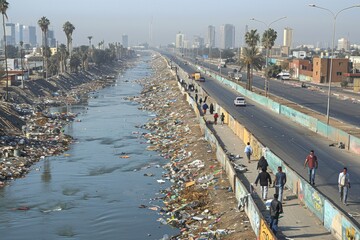 People walking along a polluted river while buildings tower in the background on a clear day, highlighting urban environmental issues