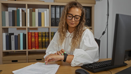 Woman checking time at the office desk with documents, wearing glasses and smart watch, surrounded by books, looking focused and professional in an indoor workplace setting