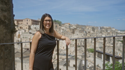 Wall Mural - Young hispanic woman in black dress poses at a scenic viewpoint overlooking the historic old town of matera, basilicata, italy on a sunny day.