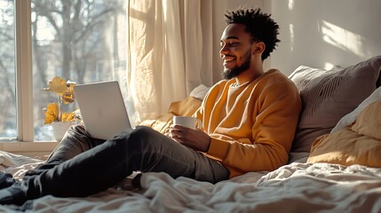 A man sitting comfortably on a bed, holding a cup while working on his laptop, smiling in a bright, sunlit winter room.