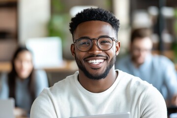 A young man with glasses and a well-groomed beard is sitting in an office environment, smiling confidently with colleagues blurred in the background, suggesting teamwork.