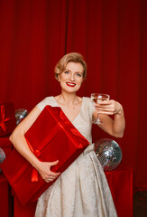 mature stylish elegant woman in cocktail dress with glass of sparkling wine with presents on red background. Happy New Year!