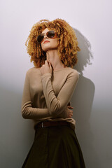 Fashionable woman with curly hair wearing sunglasses, posing confidently against a minimalist light background, showcasing a contemporary style and elegance with a natural vibe