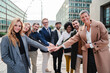 © Jose Calsina - Large group of diverse business people standing outdoors, smiling, and stacking hands together, showing teamwork, unity, partnership, confidence and collaboration in a modern office district setting