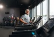© YiuCheung - A senior man with grey hair and beard runs on a treadmill in a gym.