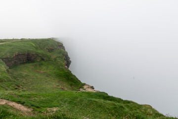 Cliffs of Moher in County Clare, Ireland, reveals a grassy cliff edge fading into the dense mist. The soft light and overcast conditions create a peaceful yet mysterious atmosphere