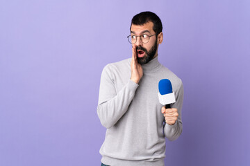 Adult reporter man with beard holding a microphone over isolated purple background whispering something with surprise gesture while looking to the side