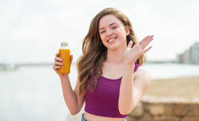 Wall Mural - Young redhead girl holding an orange juice at outdoors saluting with hand with happy expression