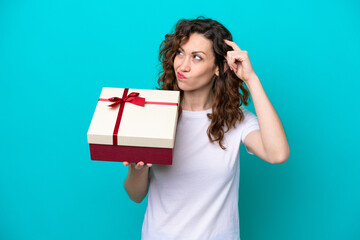 Wall Mural - Young caucasian woman holding a gift isolated on blue background having doubts and with confuse face expression