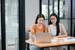 © Satori Studio - Two women working together at a desk in a contemporary office, using a laptop and reviewing documents.