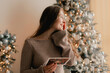 © Forewer - Side view of charming woman reading Christmas greeting card on background of Xmas tree at cozy living room with festive interior. Surprised lady pleasure reading letter at home. Part of a series