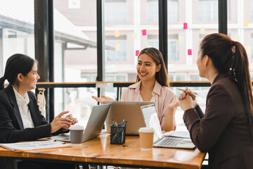 Wall Mural - Efficient teamwork is showcased in this modern office setting, where three women engage in productive discussion around table filled with laptops and coffee cups. Their expressions reflect