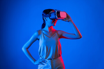 Smart female standing hit by neon light wearing VR headset connecting metaverse, futuristic cyberspace community technology. Elegant woman confidently pose while look into virtual area. Hallucination.