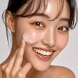 © Un_Un - Beautiful young woman with clear skin applying moisturizer cream on her face with fingers, smiling and looking at camera, facial beauty and skincare concept. Generative AI
