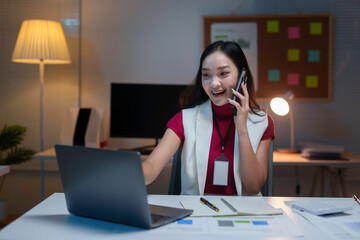 Young professional working late in the office, talking on her phone and smiling at her laptop