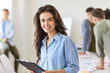 © Studio Romantic - Portrait of young smiling Caucasian girl standing on workplace in office with clipboard. Joyful woman wearing light blue shirt looking at camera with team of colleagues working on blurred background.