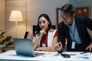 Two asian business people working late together in office using laptop computer and analyzing financial chart