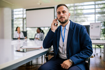 Portrait of young businessman sitting in his office in the middle of the meeting talking on the phone.