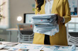©  NCST Studio - Stressed businesswoman in a busy office with a huge stack of paperwork, showing the challenges of managing documents in a corporate setting