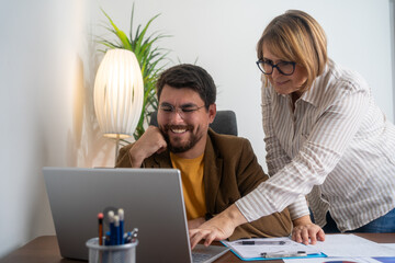 Two smiling professional business people talking using laptop computer working in office. Happy colleagues or entrepreneurs team man and woman discussing corporate technology at workplace.