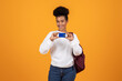 © Prostock-studio - A young woman stands with a backpack, cheerfully holding a smartphone. She wears a cozy sweater and blue jeans, radiating positivity against a vibrant yellow background.