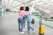 © Prostock-studio - Three friends share a heartfelt hug at a bustling airport terminal. One is wearing a pink sweater, and the others are in a light blue outfit, with a yellow suitcase nearby, ready for their journey.
