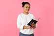 © Prostock-studio - A young Asian woman stands joyfully reading a book, her expression vibrant and engaged. The backdrop is a solid bright pink, enhancing her cheerful demeanor.