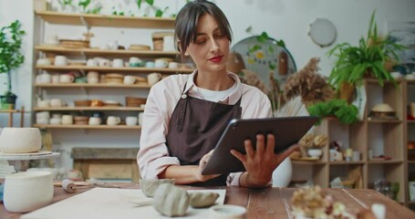 Camera moving around attractive Caucasian woman. Girl working as potter in studio. Looking at her tablet screen. Typing with fingers on touch display. Browsing social media. Searching for inspiration.