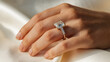 © Hound - A woman is holding her hand up to show off her diamond ring. The ring is large and has a diamond in the center. The woman's hand is on a white cloth, which adds to the elegance of the scene