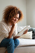 © Daniel - Vertical portrait of young happy Hispanic woman using digital tablet sitting on the sofa at home living room.