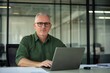 © 为轩 张 - Middle-aged man with glasses working on a laptop at an office desk, blurred glass wall background, wide-angle professional photograph.