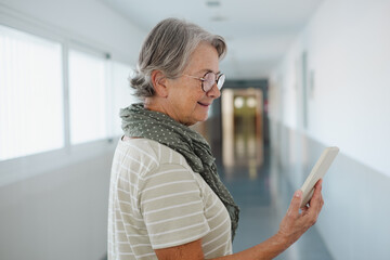 Back view of senior Caucasian woman walking in a corridor of a building while using the phone. Smiling elderly lady reading a message on smartphone