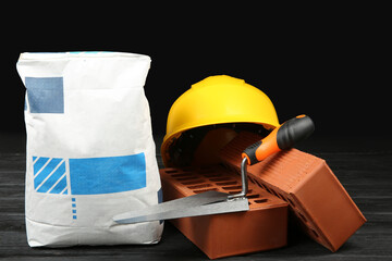 Canvas Print - Red bricks, bag of cement powder, putty knife and yellow hard hat on dark wooden table. Building materials