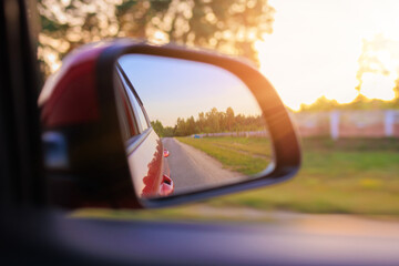 The sun is reflected in the mirror, evening road. View in the side rear view mirror of a red car driving along the highway