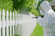 © Kanchanit - Chemical Spraying by Pest Control Expert in White Hazmat Suit by Suburban White Fence and Lush Green Garden