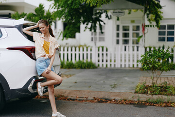 Stylish young woman leaning against a modern white car, dressed in casual attire with a relaxed pose Scenic street with greenery and a white fence in the background creates a vibrant atmosphere