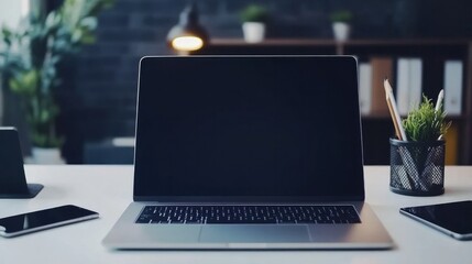 close-up of a laptop open on a white surface. A tablet PC, a smartphone, pencil holders, and paper graphics are all close by. Houseplants and a shadowy wall are in the backdrop. Workplace empty.