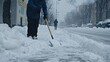 © lililia - On a snowy day, a man diligently shovels snow off the sidewalk in a city street as vehicles drive past