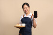 © PBXStudio - Asian female waitress in apron holding bread and coffee drink on wooden tray with smartphone on beige background.