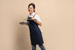 © PBXStudio - Beautiful and attractive Asian female coffee shop owner wearing apron. She holds a frying pan and a wooden spatula on a beige background.