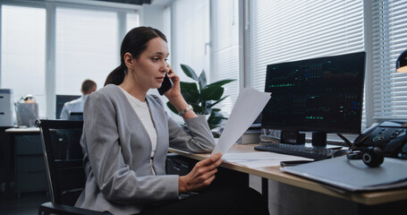 Female financial analyst discusses strategy of project with colleague using mobile phone. Specialist from marketing department checks data in documents with parameters displayed on PC monitor.