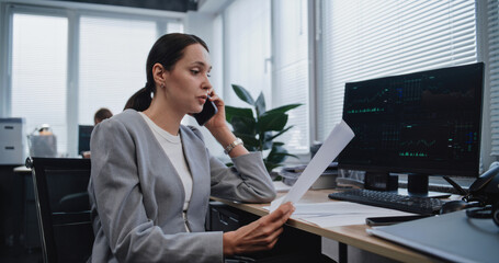 Female financial analyst discusses strategy of project with colleague using mobile phone. Specialist from marketing department checks data in documents with parameters displayed on PC monitor.