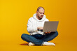 © Davidovici - Young serious caucasian man sitting on floor and using modern notebook, typing on laptop, isolated on yellow background with empty space for text