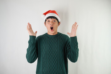 Surprised young asian man wearing santa clause hat and green long sleeved sweater win a prize or good news, isolated over white background. Concept for Christmas Holiday and New Year Party