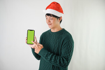 Excited young asian man wearing santa clause hat and green long sleeved sweater is showing smartphone green screen, isolated over white background. Concept for Christmas Holiday and New Year Party