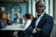 © N Joy Art  - Mature African American businessman in a navy blazer standing confidently with arms crossed in a modern boardroom. Meeting in progress in background with colleagues seated, demonstrating leadership